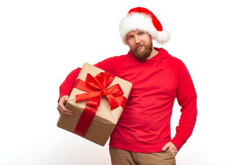 A brutal man with a red beard in a santa claus hat looks at the camera and holds a gift box with a red ribbon in his hands. Studio shot isolate, christmas concept.