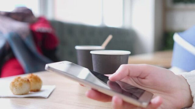 Pone in hands on the background of a cup of coffee. The girl is texting on her smartphone. There is a cup of coffee on the table. She is sitting in a cafe