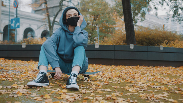 Young hispter woman with face mask and hoodie sitting on the skateboard in the city park in autumn. High quality photo