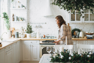 Attractive smiling woman with curly hair in plaid shirt bakes cookies at bright kitchen at the home