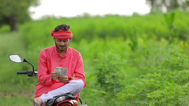 Happy Young indian farmer sitting two wheeler and counting money at field