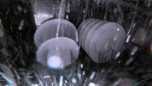 Water sprayer jets shoot water to rinse white stylish plates at dishwasher operating close inside view through glass wall