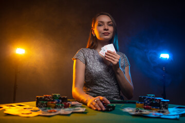 Woman holds her card and sits at poker table in casino on dark smoky background with lamps light. Unemotional face. Gambling concept.