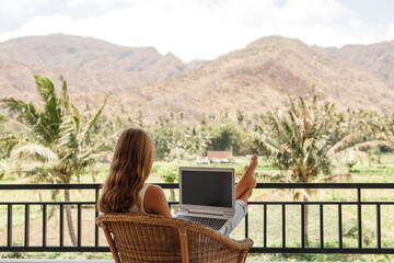 Back view young woman with laptop sit on wicker  chair on the balcony,  freelancer at work. Tropical nature view