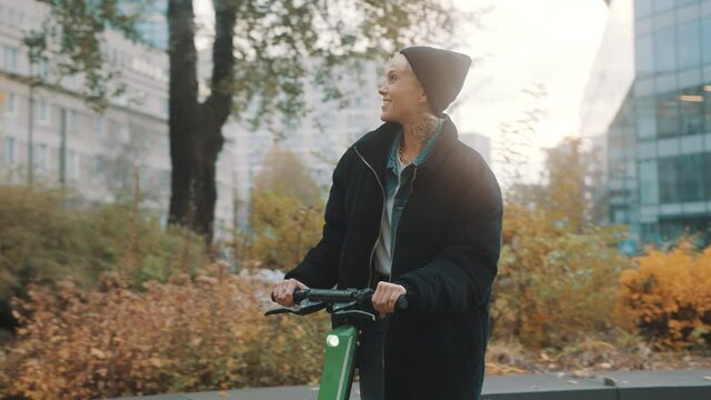 Young woman in winter black jacket with hat and face mask riding electic scooter in the city in autumn. High quality 4k footage
