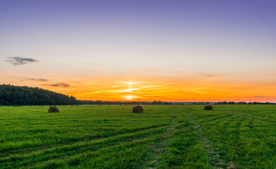 Naklejka na ściany i meble Scenic view at beautiful sunset in green shiny field with hay stacks, bright cloudy sky, country road and golden sun rays with glow, summer valley landscape