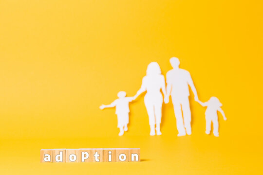 A Family Cut Out Of White Paper On A Yellow Background. Wooden Cubes With The Inscription Adoption. Copy Space. The Concept Of Adoption