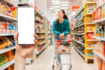 A person's hand holds a mobile phone, with a shopping basket on the screen. Mock up. In the background, a woman is pushing a grocery cart in a supermarket, in a blur. The concept of online shopping