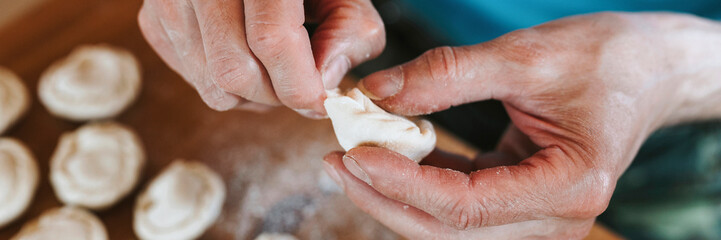 Fototapeta premium hands of senior man cooking and molding small homemade uncooked dumplings with meat on kitchen table. national traditional Russian cuisine. do it yourself. top view. banner