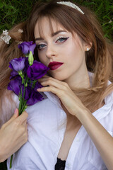 A beautiful, young girl lies on green grass in a white shirt with purple flowers in her hands. Close-up portrait.