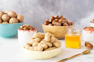 Different nuts in beautiful wooden and ceramic bowls (cashews, hazelnuts, walnuts) stand on a white table against a gray wall. There is a small jar of honey on the right. Healthy organic food