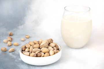 A glass of very healthy pistachio milk stands on a gray-white concrete background. There are pistachio nuts in a white bowl nearby. Diet milk, vegetarian food. Gluten free. Close-up.