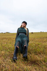 Naklejka na ściany i meble A girl with blonde hair , wearing a black cap, green chiffon dress and rough boots sits in a mown field.