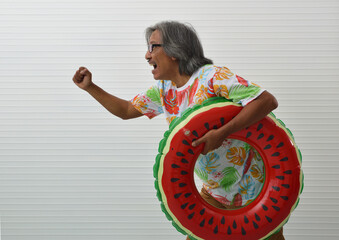 Happy elderly traveler asian man wearing summer shirt and eyeglasses holding watermelon inflatable ring running over white wall background, Business summer holiday concept