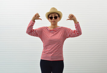 Naklejka premium Moody young asian woman wearing red stripped shirt, sunglasses, straw hat and jeans showing dislike thumps down hand sign standing over white wall background, Upset facial expression