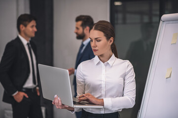 Brown-haired female standing near flipchart, checking laptop, male colleagues discussing behind