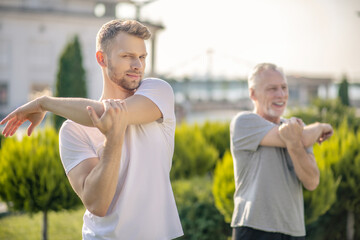 Young bearded male and mature grey-haired male stretching arms outside