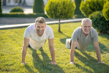 Young male and grey-haired male doing push ups on grass, smiling