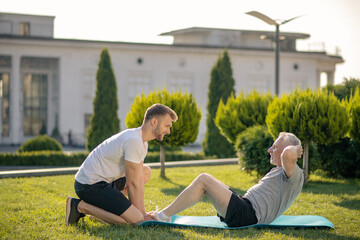 Young brown-haired male helping mature grey-haired male with sit ups - obrazy, fototapety, plakaty