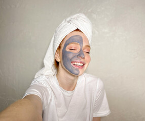 woman with a smile and a clay mask on half of her face takes a selfie on a smartphone at home. enjoying beauty treatments.