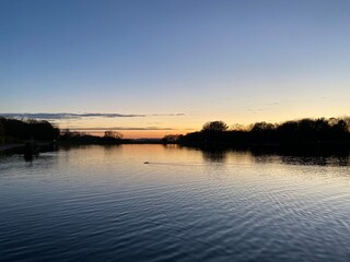 Wiew at sunset with blue sky with some clouds at the horizon reflected on a lake in a cold autumn day, intnse orange and dark colours at dusk in Stevenage, Hertfordshire, United Kingdom