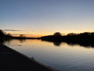 View at sunset with blue sky with some clouds at the horizon reflected on a lake in a cold autumn day, intense orange and dark colors at dusk in Stevenage, Hertfordshire, United Kingdom
