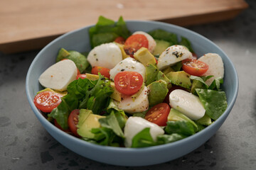 Salad with avocado, cherry tomatoes, romaine and mozzarella in blue bowl on concrete countertop