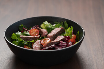 Steak salad with cherry tomatoes and mixed greens in black bowl on walnut wood background