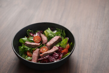 Steak salad with cherry tomatoes and mixed greens in black bowl on walnut wood background