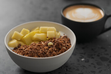 chocolate granola with mango and greek yogurt in white bowl with black cup with cappuccino on concrete background