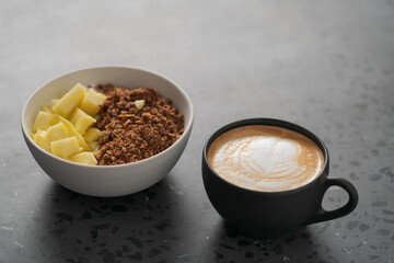 chocolate granola with mango and greek yogurt in white bowl with black cup with cappuccino on concrete background