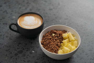 chocolate granola with mango and greek yogurt in white bowl with black cup with cappuccino on concrete background
