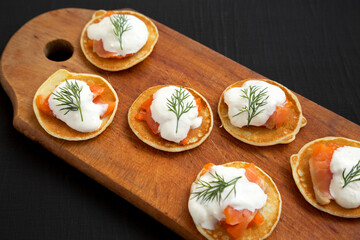 Homemade Blini with Smoked Salmon, Creme and Dill on a rustic wooden board on a black surface, side view. Close-up.
