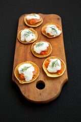 Homemade Blini with Smoked Salmon, Creme and Dill on a rustic wooden board on a black background, low angle view.