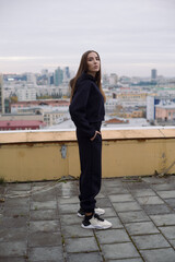 Full height photo of young woman, standing on roof of tall building in large city.