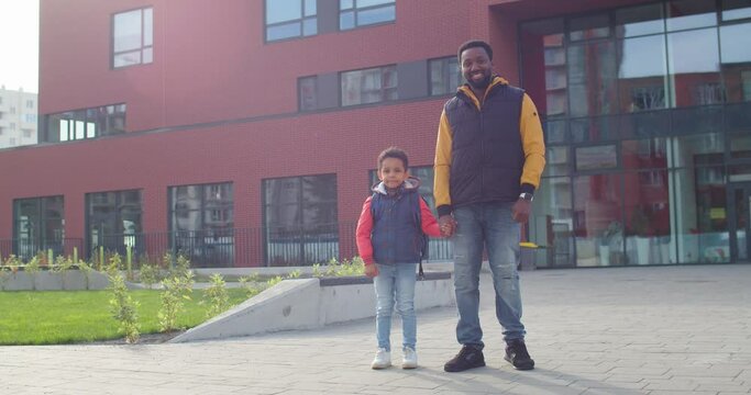Cheerful African American man standing outdoors near school with cute little son pupil before classes Happy father bringing small junior student to school and smiling. Education concept Back to school