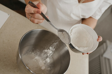 A close photo of the falling wheat flour from the spoon in the hand of the young woman into the stainless steel bowl of the mixer.