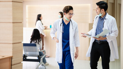 Fototapeta premium Asian Doctors talking and discussing about patient's case while walking through corridor in hospital. Two young Asian men medical practitioners talking and consulting together.