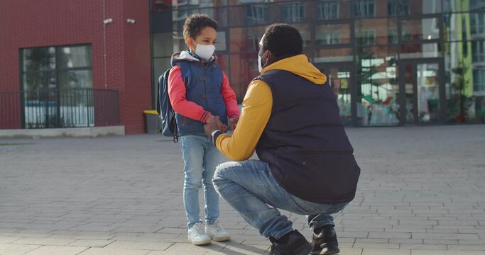 Young male talking and cheering up little cute African American schoolboy in mask with backpack and preparing for first school day Boy pupil speaking with father outdoors Back to school Family concept