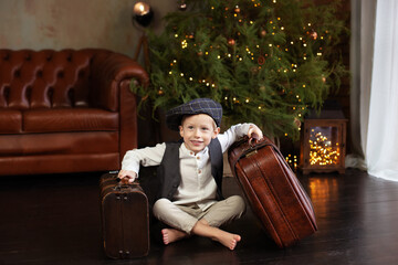 Little boy sit on wooden floor with christmas suitcase in Cozy living room. Smiling Little blond boy in white shirt with vest and in cap sit by Christmas tree with old suitcases. Loft interior Xmas