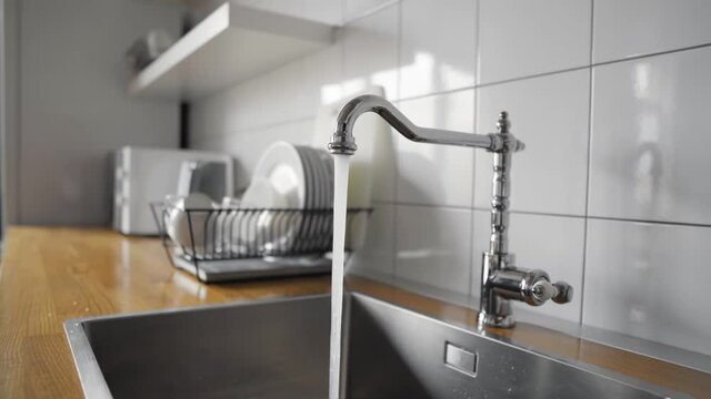Water flowing out of a kitchen stainless steel tap into the sink. Wasting water by leaving a chrome faucet tap running. Water misuse in domestic duties and activities. Overusing household water.
