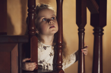 A cute little girl sits on a wooden staircase and stuck her head through the railing. Low key. Soft focus. Inside