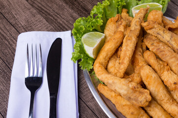 Portion and fried fish served on lettuce leaves with lemons and tartar sauce to accompany. Fork and knife on white napkin. Gastronomic photography close up. Popular restaurants.