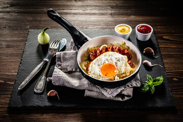 Fried sausages with egg and onion in frying pan on wooden table
