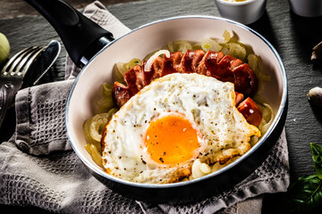 Fried sausages with egg and onion in frying pan on wooden table

