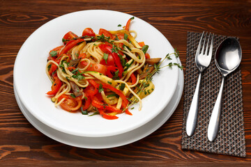 Vegetarian pasta with paprika and tomatoes on a white plate on a dark wooden table, fork and spoon