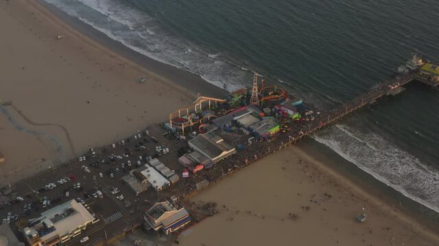 Santa Monica Pier, Los Angeles from above at beautiful golden hour Sunset packed with Tourists, Pedestrians having fun at theme park Ferris Wheel with ocean view, Aerial Wide View, Dolly slide right