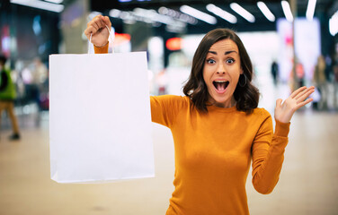 Close up photo of a cute young woman is showing white showing bag in the camera on the mall...