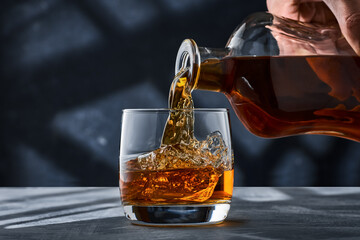 Round glass of whiskey with ice on a concrete table, a glass is filled with whiskey from a bottle.