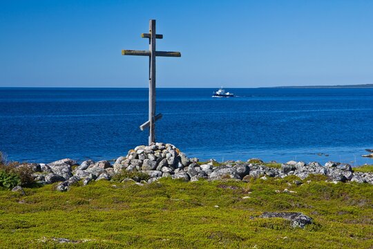 The wooden cross on a grave on the grassy shore of the White Sea and a ship in the background in summer on Solovetsky Islands, Russia.